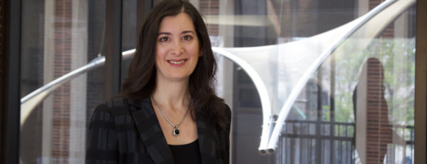 Professor Nancy Rothbard has brown hair, and is wearing a black and gray plaid jacket and a black necklace. She is smiling.