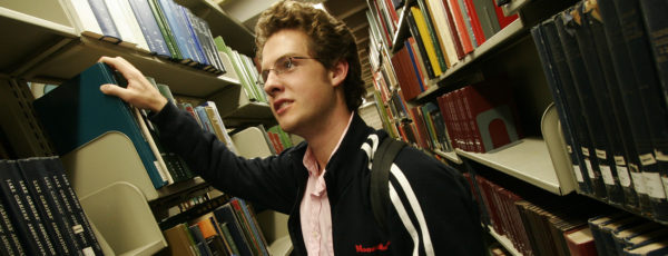 A student wearing casual clothes stands in the stacks of Van Pelt Library. He has blonde hair and blue eyes. He is wearing glasses.