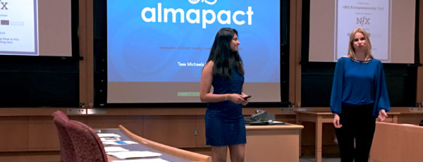 Two women present in front of a classroom. One wears a blue dress, the other a blue shirt with three screens behind them.