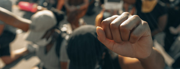 A raised fist in the foreground, with people kneeling and sitting in the background, suggesting a protest or demonstration for solidarity or social justice.