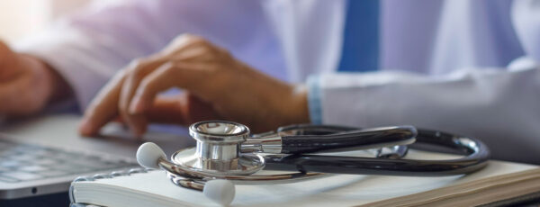 A person in a white coat typing on a laptop, with a stethoscope resting on an open notebook. Represents medical work or healthcare technology.