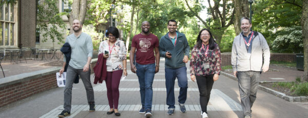 A group of six people is walking together on a tree-lined pathway, smiling and appearing to enjoy a casual outing.