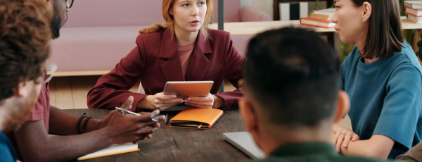 A group of people having a meeting around a table, with one person speaking and holding a tablet. Various notebooks and laptops are visible.