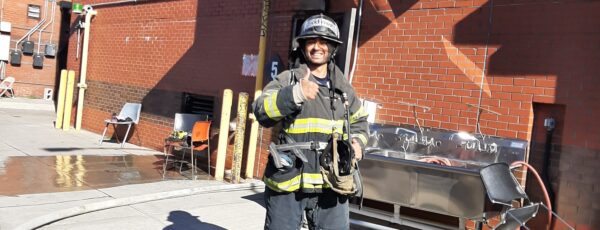 A firefighter in full gear stands outdoors, giving a thumbs-up. They''re in front of a brick building with yellow barriers and equipment nearby.