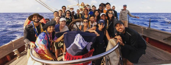 A diverse group of people smiling on a sailing boat holding a flag. The ocean is visible in the background, suggesting a group adventure or expedition at sea.