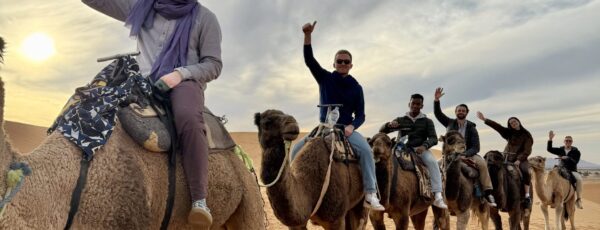 A group of people riding camels in a desert setting, waving and smiling. The sky is cloudy with a sun near the horizon, creating a warm and adventurous atmosphere.