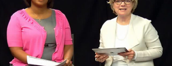Two women, one in pink, one in white, sitting in front of a black background and holding papers as they speak to the camera.